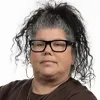 Portrait of a woman with curly black and gray hair tied up, wearing black glasses and a dark top, facing the camera against a white background.
