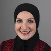 Portrait of a smiling woman wearing a black hijab and a black and red patterned top, posed in front of a gray background.