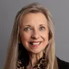 Professional headshot of a woman with long straight blonde hair, wearing a black outfit with a patterned scarf, smiling warmly in front of a gray background.