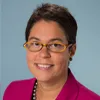 Portrait of a woman with short dark hair wearing yellow glasses, a magenta blazer, and a pearl necklace, posed in front of a light blue background.