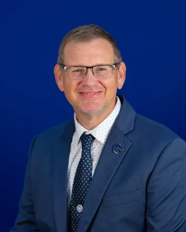 Professional headshot of a man wearing glasses, a blue suit jacket, white patterned shirt, and blue tie, smiling in front of a solid blue background.