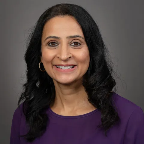 A woman with long dark hair wearing a purple blouse smiles in front of a gray background, posing for a professional headshot.