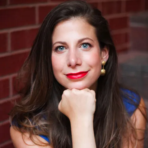 Professional headshot of a woman with long brown hair and bright blue eyes, wearing a sleeveless blue top and gold earrings, resting her chin on her hand and smiling slightly against a brick wall background.