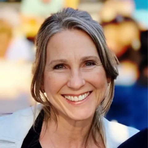 Close-up headshot of a smiling woman with long gray hair parted to the side, wearing a light-colored jacket, photographed outdoors with a softly blurred background of people and warm-toned surroundings.