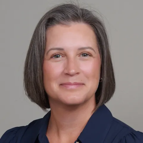 Professional headshot of a woman with short gray-brown hair, wearing a navy blouse, smiling softly against a neutral gray background.