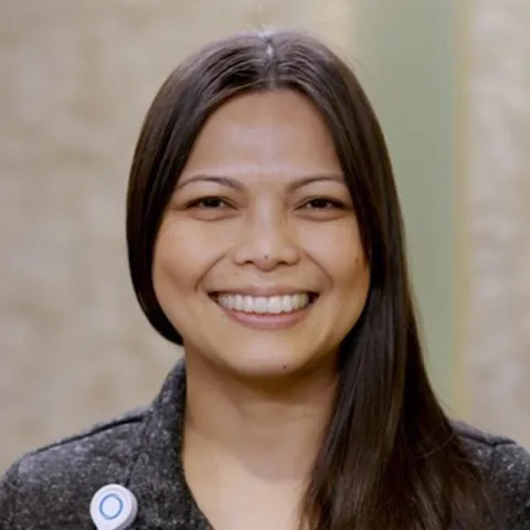 Professional headshot of Marie Yabut, smiling, with long dark hair worn to one side, wearing a dark jacket and an ENA pin, photographed against a neutral indoor background.