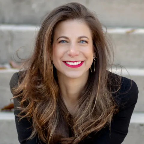 Professional headshot of Rachel Tenenbaum, smiling, with long brown hair worn loose, bright red lipstick, and gold hoop earrings, wearing a dark blazer, seated on outdoor stone steps with a softly blurred background.