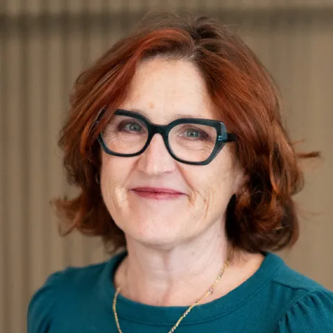 Professional headshot of a woman with short auburn hair and black-framed glasses, wearing a teal top and gold necklace, smiling softly against a neutral indoor background.