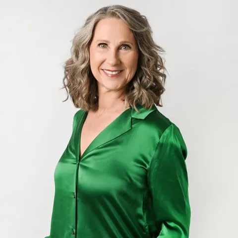 Professional portrait of a woman with shoulder-length curly gray hair wearing a green satin blouse and white pants, standing against a white background with a relaxed smile and hands in her pockets.
