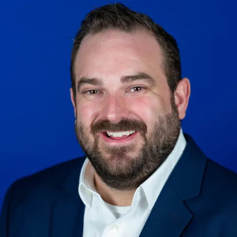 Professional headshot of a smiling man with short dark hair and a full beard, wearing a navy blazer and white collared shirt against a solid blue background.