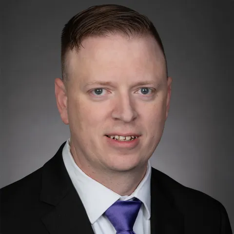 Professional headshot of a man wearing a dark suit jacket, white shirt, and purple tie, with neatly styled hair, facing the camera against a neutral gray background.