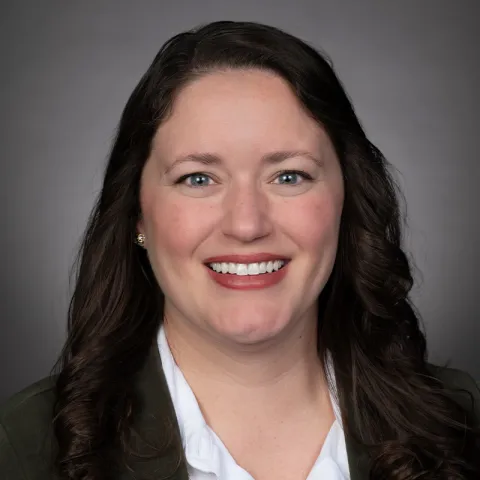Professional headshot of a woman with long, dark wavy hair, wearing a white blouse and dark jacket, smiling against a neutral gray background.