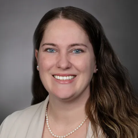 Professional headshot of a woman with long, dark brown hair, wearing a light-colored blazer and pearl necklace, smiling against a neutral gray background.