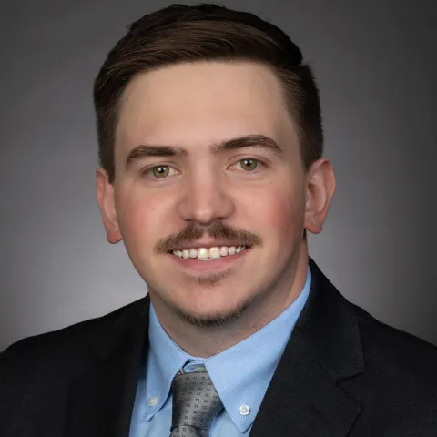Professional headshot of a man with short brown hair and a trimmed mustache, wearing a dark suit jacket, light blue shirt, and gray tie, smiling against a neutral gray background.