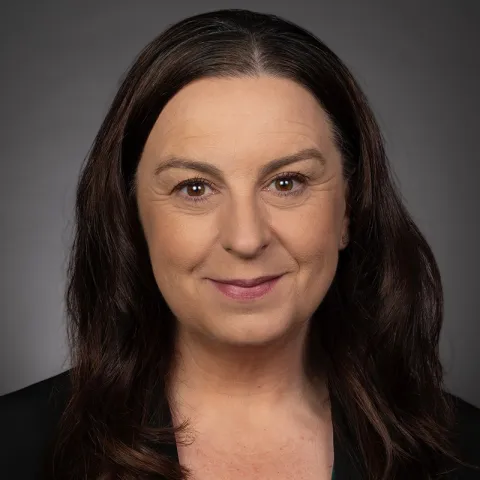 Professional headshot of a person with long dark hair and a subtle smile, wearing a dark blazer, photographed against a neutral gray background.